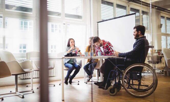 homem em cadeira de rodas sorrindo enquanto participa de uma reunião colaborativa com colegas em um escritório moderno e iluminado representando o que é pcd no contexto de inclusão profissional e acessibilidade corporativa