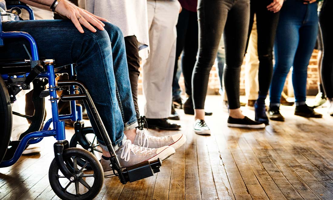 pessoa em cadeira de rodas azul integrada a um grupo de colegas de trabalho em pé sobre piso de madeira representando a prática de recrutamento inclusivo no ambiente corporativo.
