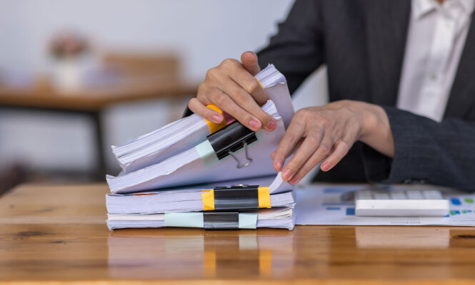 pessoa em traje social organizando uma pilha de papeis com clipes em uma mesa de madeira representando a separacao de documentos do rh durante uma fiscalizacao da lei de cotas.