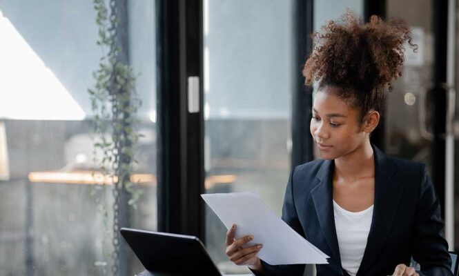 uma mulher negra com roupas sociais analisa atentamente documentos impressos em uma mesa de escritorio ilustrando a rotina do rh ao validar o certificado de reabilitação profissional de um candidato