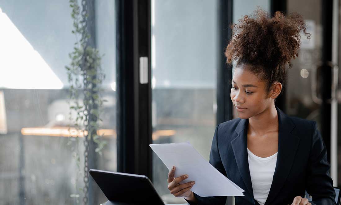 uma mulher negra com roupas sociais analisa atentamente documentos impressos em uma mesa de escritorio ilustrando a rotina do rh ao validar o certificado de reabilitação profissional de um candidato